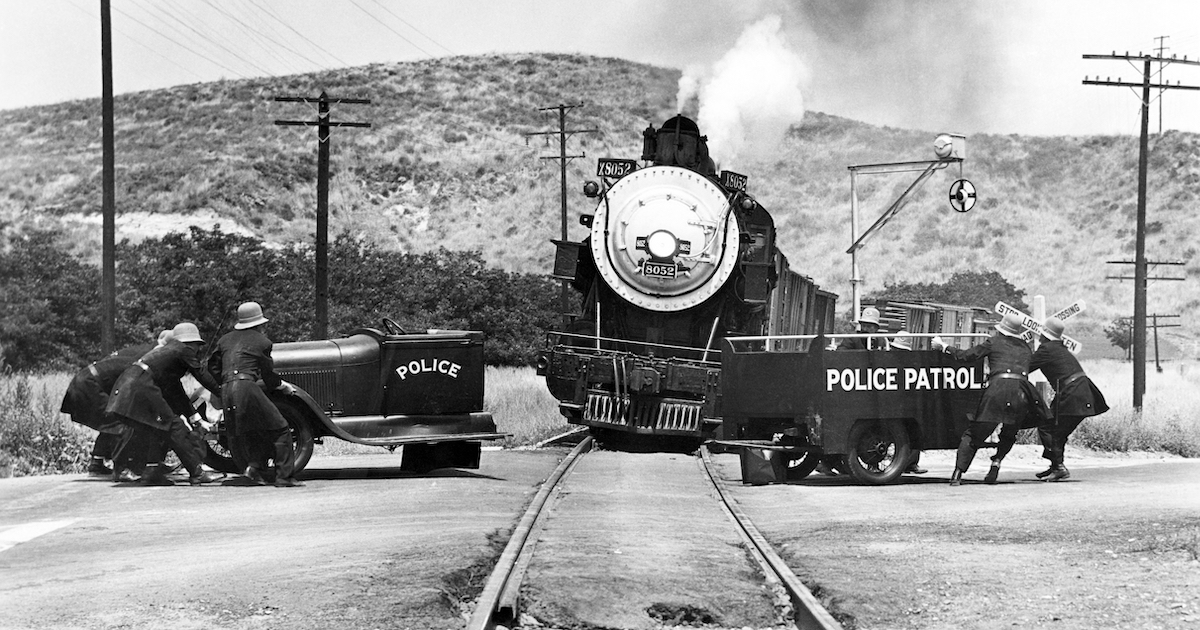 A train coming down the tracks towards the viewers, with a police vehicle split in two, with one part on either side of the tracks.