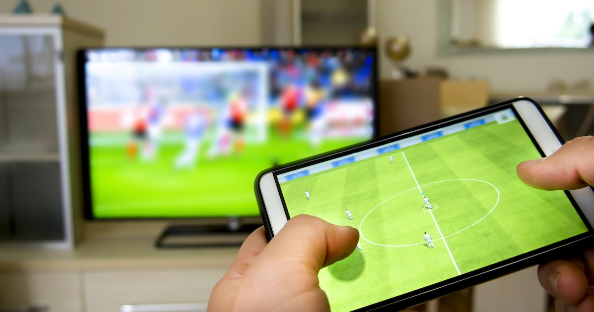 A soccer game on TV in the background with hands holding a tablet in the foreground